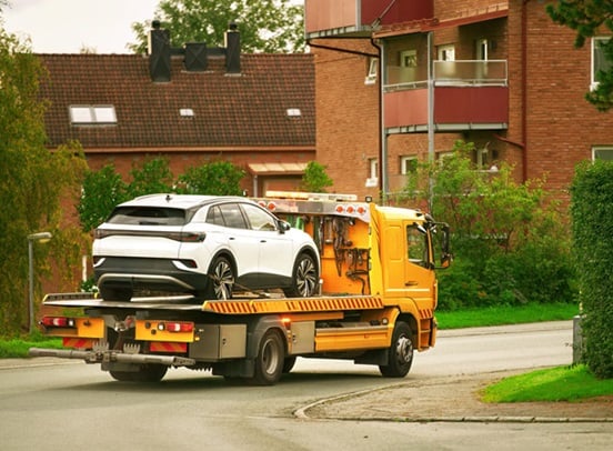 Yellow recovery truck carries white car through British suburb